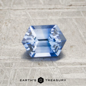 A 0.95-carat heated blue sapphire with a hexagonal, faceted cut rests on a textured, light surface above the "EARTH'S TREASURY" logo.
