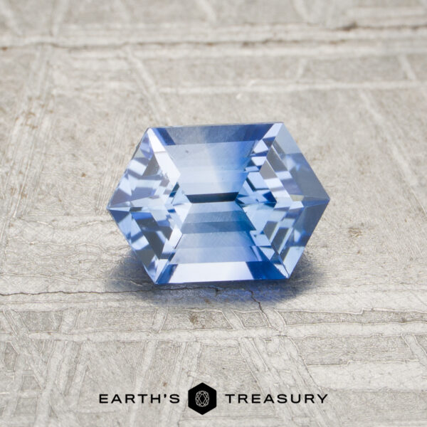 0.95-Carat Blue-Colorless Particolored Ceylon Sapphire (Heated) A 0.95-carat heated blue sapphire with a hexagonal, faceted cut rests on a textured, light surface above the "EARTH'S TREASURY" logo.