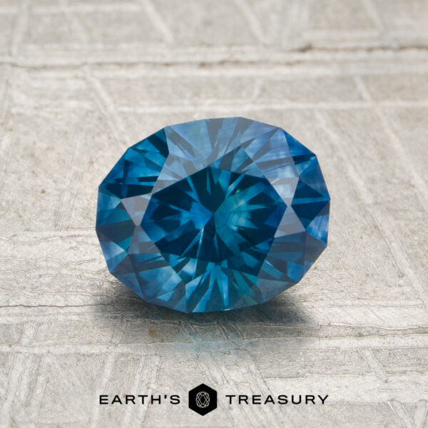 1.61-Carat Rich Teal Montana Sapphire (Heated) A 1.61-carat heated Montana sapphire, faceted in an oval shape, rests on a textured light gray surface with "EARTH'S TREASURY" beneath.