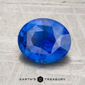 A 2.03-carat heated blue sapphire with an oval cut rests on a textured gray surface, displayed above the Earth's Treasury logo and name.