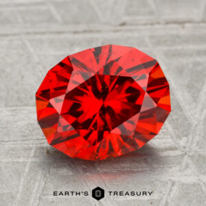 A 3.47-carat spessartine garnet, round and faceted, is displayed on a textured gray surface with "EARTH'S TREASURY" shown at the bottom.