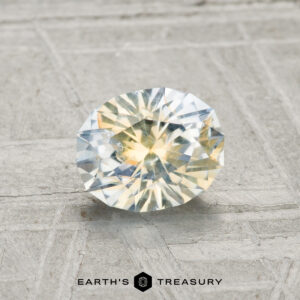 A 0.96-carat Montana Sapphire with yellow and blue hues rests on a textured gray surface, with the "EARTH'S TREASURY" logo at the bottom center.