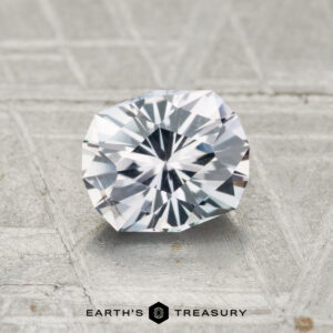 A 1.04-carat round brilliant Montana sapphire with angular facets rests on a textured gray surface; the "Earth's Treasury" logo is shown at the bottom.