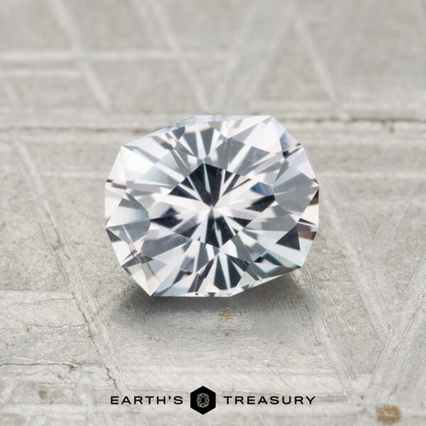 1.04-Carat Light Gray to Lilac Color-Change Montana Sapphire A 1.04-carat round brilliant Montana sapphire with angular facets rests on a textured gray surface; the "Earth's Treasury" logo is shown at the bottom.