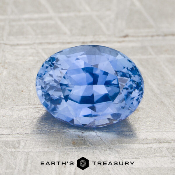 2.02-carat heated sapphire, oval-cut and blue, displayed on a textured gray surface with "EARTH'S TREASURY" text and logo beneath.