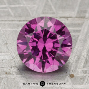 A 3.12-carat pastel pyrope garnet cut in a round brilliant style sits on a textured gray surface, with the text "EARTH'S TREASURY" displayed below.