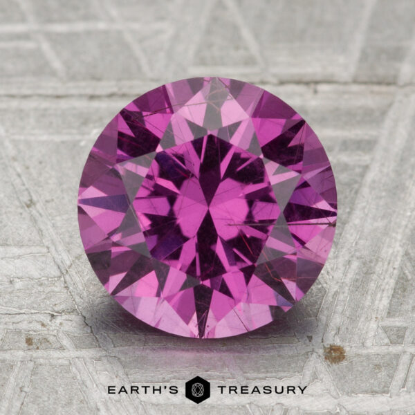 3.12-Carat Pastel Pyrope Garnet A 3.12-carat pastel pyrope garnet cut in a round brilliant style sits on a textured gray surface, with the text "EARTH'S TREASURY" displayed below.