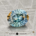 A 2.15-carat Montana sapphire with blue and yellow hues rests on a textured gray surface, with the text "EARTH'S TREASURY" below.