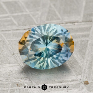 A 2.15-carat Montana sapphire with blue and yellow hues rests on a textured gray surface, with the text "EARTH'S TREASURY" below.