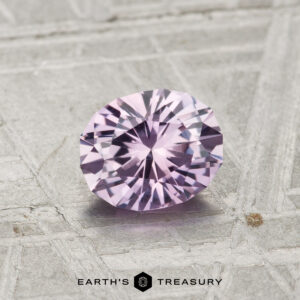 A 0.96-carat Montana sapphire, faceted in an oval cut and displaying pale purple hues, rests on a textured grey surface with the Earth's Treasury logo below.