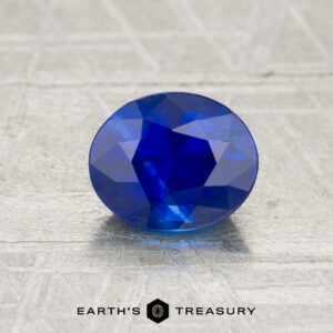 A 1.18-carat oval-cut blue sapphire displayed on a textured surface, with "EARTH’S TREASURY" and a hexagonal logo beneath.