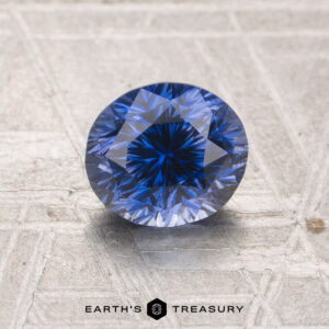 A 1.30-carat unheated Ceylon sapphire, round brilliant-cut, rests on a textured metallic surface with the "Earth's Treasury" logo and text below.