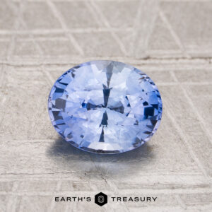 A 1.68-carat heated Ceylon sapphire, faceted in an oval shape, is shown on a textured light gray surface with "EARTH'S TREASURY" displayed at the bottom.