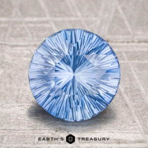 A round, brilliantly-cut 2.68-carat heated Ceylon sapphire is displayed on a textured surface with "Earth’s Treasury" written below it.