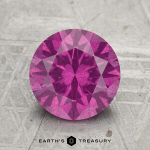 A round, faceted 2.50-carat pastel pyrope garnet is displayed on a textured surface with "EARTH'S TREASURY" written below.