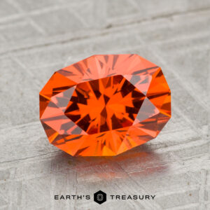 A 2.74-carat Spessartine Garnet, oval-cut and faceted, is showcased on a textured gray surface with the "EARTH'S TREASURY" text and logo beneath it.