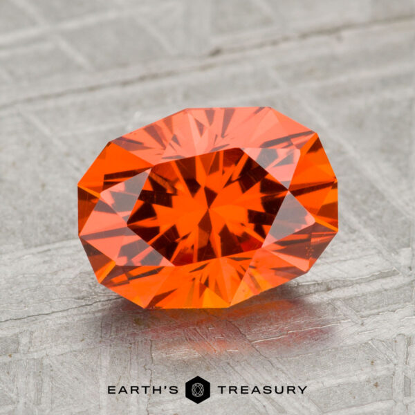 2.74-Carat Spessartine Garnet A 2.74-carat Spessartine Garnet, oval-cut and faceted, is showcased on a textured gray surface with the "EARTH'S TREASURY" text and logo beneath it.