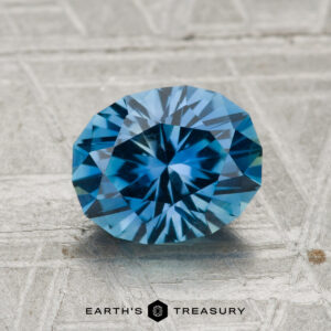 A 1.95-carat Montana Sapphire, round brilliant-cut and blue, is displayed on a textured gray surface with "Earth's Treasury" below it.