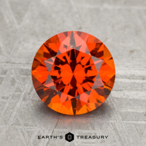 A 2.57-carat Spessartine Garnet, round and faceted with an orange-red hue, is showcased on a textured gray surface featuring the text "EARTH'S TREASURY" at the bottom.