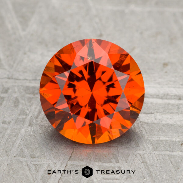 2.57-Carat Spessartine Garnet A 2.57-carat Spessartine Garnet, round and faceted with an orange-red hue, is showcased on a textured gray surface featuring the text "EARTH'S TREASURY" at the bottom.