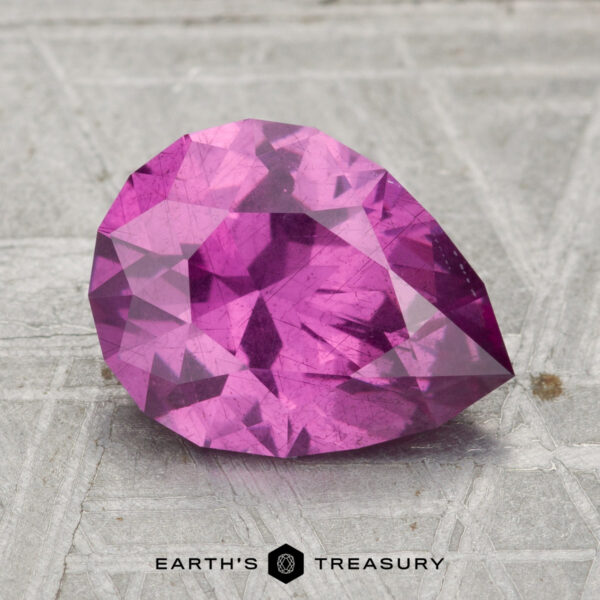 A 2.43-carat pastel pyrope garnet, cut in a faceted pear shape, rests on a textured grey surface with "EARTH'S TREASURY" displayed below.