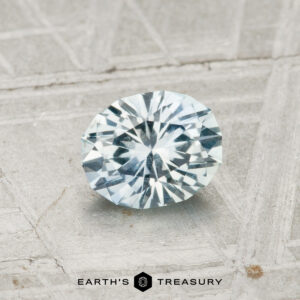 0.84-carat Montana Sapphire, oval-cut with a light bluish hue, displayed on a textured gray surface, "Earth's Treasury" logo and text at the bottom.