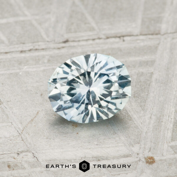 0.84-Carat Montana Sapphire 0.84-carat Montana Sapphire, oval-cut with a light bluish hue, displayed on a textured gray surface, "Earth's Treasury" logo and text at the bottom.