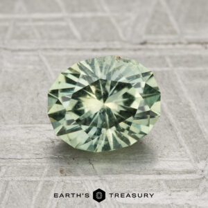 A round, faceted light green 1.79-carat Montana sapphire is displayed on a textured gray surface with "EARTH'S TREASURY" at the bottom.