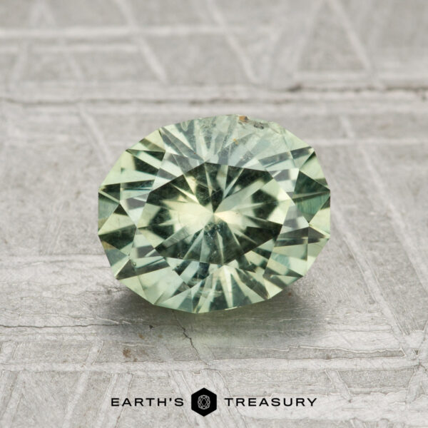 1.79-Carat Mint Green Montana Sapphire A round, faceted light green 1.79-carat Montana sapphire is displayed on a textured gray surface with "EARTH'S TREASURY" at the bottom.
