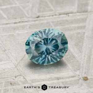 A round, faceted 0.82-carat Montana sapphire with blue-green hues rests on a gray, textured surface, with "Earth's Treasury" shown at the bottom of the image.