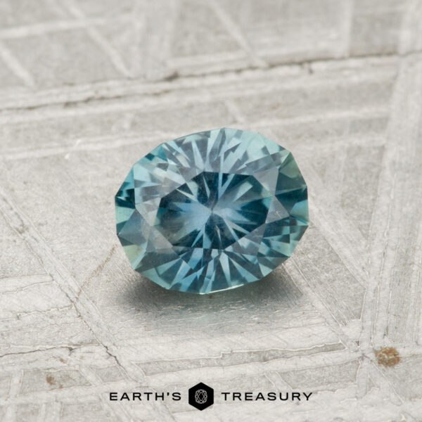 0.82-Carat Teal Montana Sapphire A round, faceted 0.82-carat Montana sapphire with blue-green hues rests on a gray, textured surface, with "Earth's Treasury" shown at the bottom of the image.