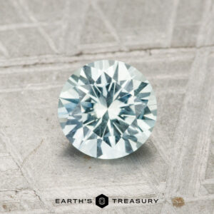 A 0.99-carat Montana sapphire, round brilliant-cut with a pale blue hue, displayed on a textured gray surface with the "EARTH'S TREASURY" logo at the bottom.