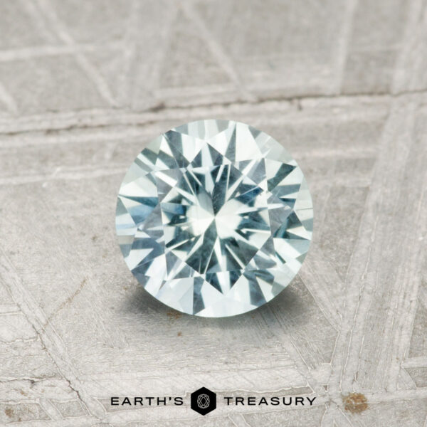 0.99-Carat Pale Aqua Green Montana Sapphire A 0.99-carat Montana sapphire, round brilliant-cut with a pale blue hue, displayed on a textured gray surface with the "EARTH'S TREASURY" logo at the bottom.