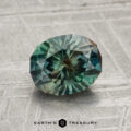 A 1.77-carat Montana sapphire, oval-cut with intricate facets and a teal-green hue, rests on a textured gray surface above the "Earth's Treasury" text.