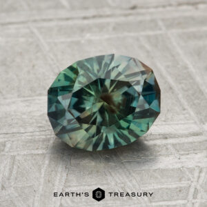 A 1.77-carat Montana sapphire, oval-cut with intricate facets and a teal-green hue, rests on a textured gray surface above the "Earth's Treasury" text.