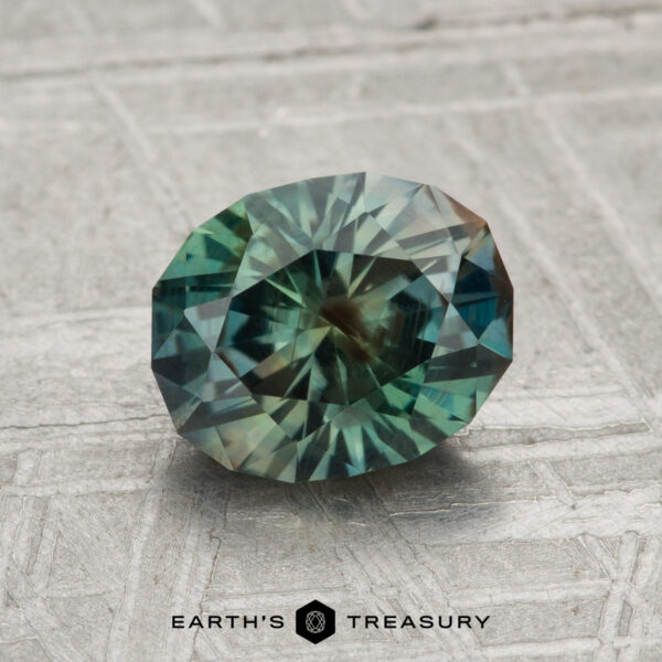 A 1.77-carat Montana sapphire, oval-cut with intricate facets and a teal-green hue, rests on a textured gray surface above the "Earth's Treasury" text.