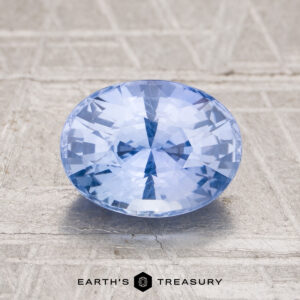 A 2.14-carat Ceylon sapphire, oval-cut and light blue, is displayed on a textured surface with the "Earth's Treasury" logo and text at the bottom.