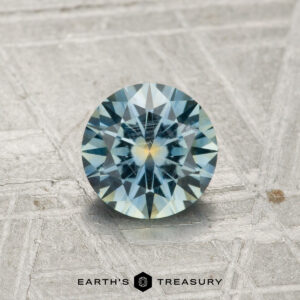 A 0.99-carat Montana sapphire with blue and yellow hues is shown on a textured gray surface, with the Earth’s Treasury logo at the bottom.
