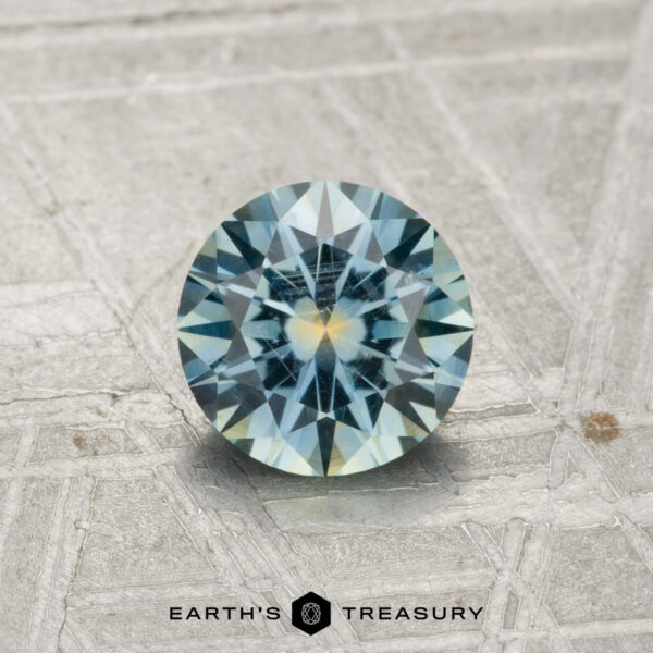 0.99-Carat Aqua Blue Montana Sapphire A 0.99-carat Montana sapphire with blue and yellow hues is shown on a textured gray surface, with the Earth’s Treasury logo at the bottom.