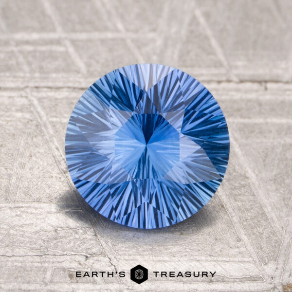 1.75-Carat Montana Sapphire (Heated) A round, faceted 1.75-carat Montana Sapphire (Heated) with a starburst pattern sits on a textured, light surface. The "EARTH'S TREASURY" logo is displayed at the bottom of the image.