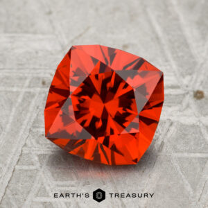 A 2.56-carat cushion-cut spessartine garnet with vivid red hues is shown on a gray, textured surface, featuring the text "EARTH'S TREASURY" at the bottom.