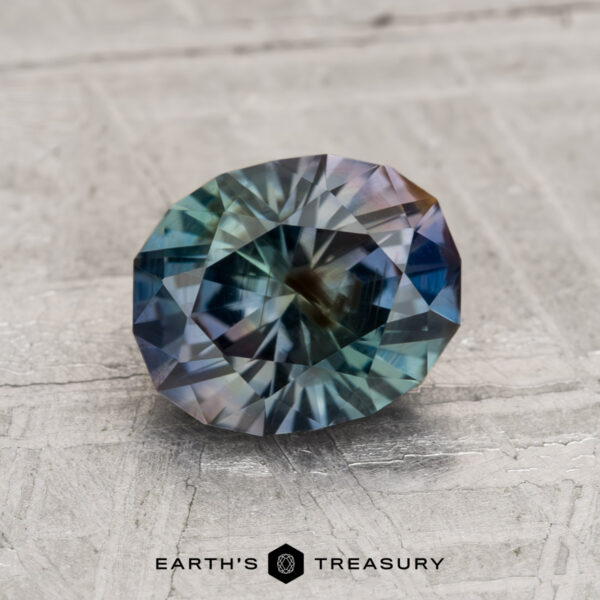 1.77-Carat Green Tricolor Color-Change Montana Sapphire A 1.77-carat Montana Sapphire with blue, green, and purple hues rests on a light textured surface, with the "EARTH'S TREASURY" logo below.