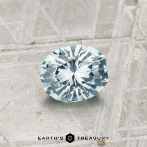A 0.82-carat round brilliant-cut Montana sapphire with a light blue hue is displayed on a textured gray surface featuring the “Earth’s Treasury” logo at the bottom.