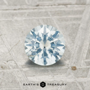A 0.94-carat Montana sapphire, round brilliant-cut with a pale blue hue, is displayed on a textured gray surface with "EARTH'S TREASURY" text below.