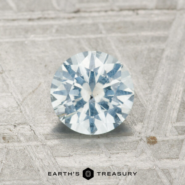 0.94-Carat Montana Sapphire A 0.94-carat Montana sapphire, round brilliant-cut with a pale blue hue, is displayed on a textured gray surface with "EARTH'S TREASURY" text below.
