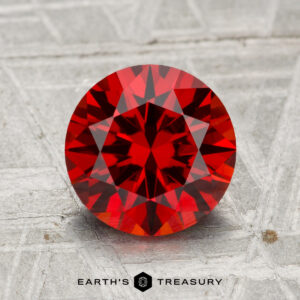 A 2.48-carat spessartine garnet, round brilliant-cut, displayed on a textured gray surface with "EARTH'S TREASURY" at the bottom.