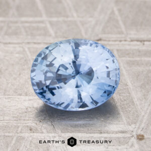 A 2.11-carat Ceylon sapphire with an oval cut and light blue hue is displayed on a textured gray surface, featuring the text "EARTH'S TREASURY" at the bottom.