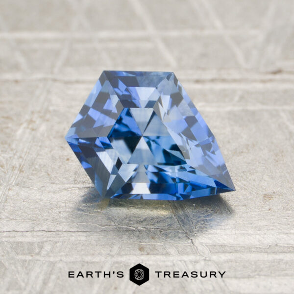 1.13-Carat Sapphire (Heated) A faceted, geometric 1.13-Carat Cornflower Blue Ceylon Sapphire (Heated) is displayed on a textured surface. “EARTH’S TREASURY” is written at the bottom.