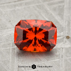 A 2.17-carat Spessartine Garnet, a faceted orange-red gemstone, is displayed on a textured gray surface with "EARTH'S TREASURY" at the bottom.