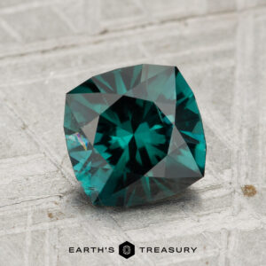 A 1.45-carat teal-blue tourmaline with a square cushion cut is displayed on a textured gray surface. "EARTH'S TREASURY" appears at the bottom of the image.